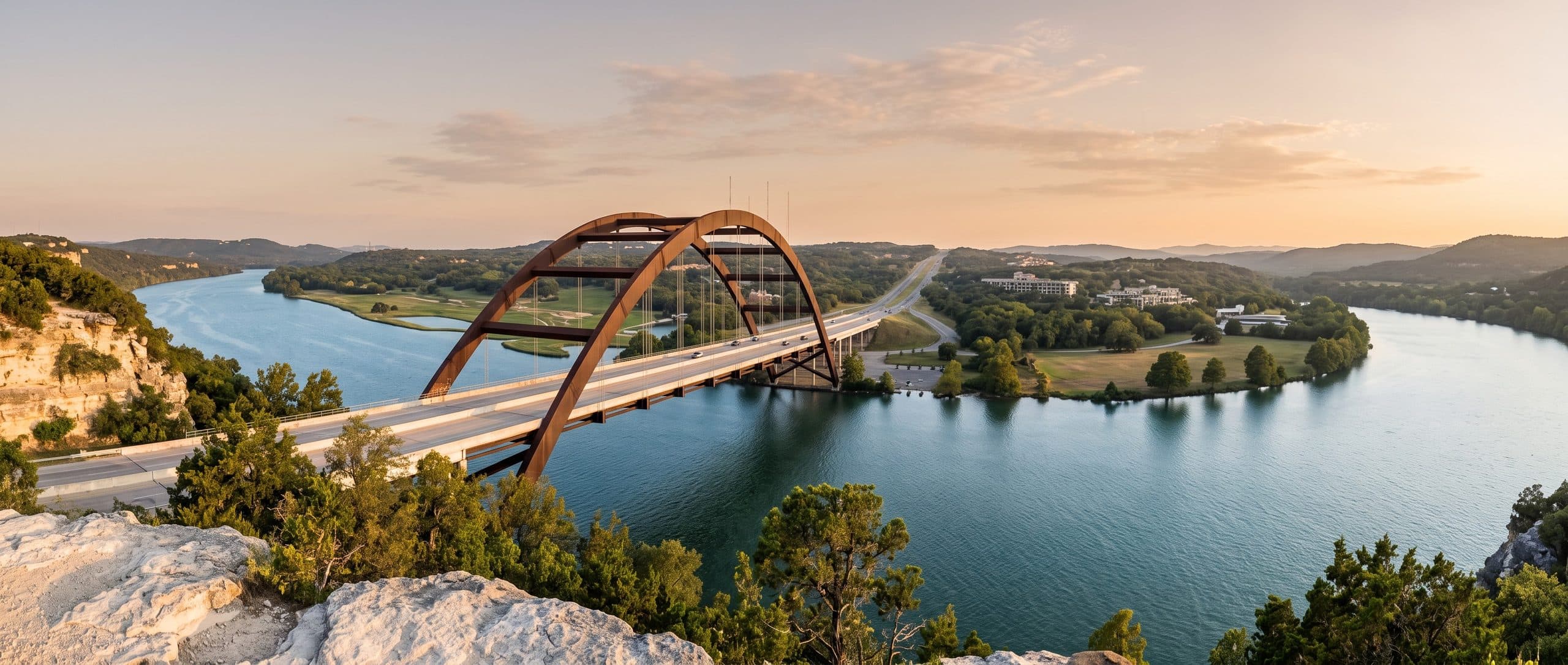 The Pennybacker Bridge in Austin seen from a clifftop at late afternoon, its rust-brown steel arch carrying a highway across a broad blue-green stretch of the Colorado River. Limestone bluffs and cedar scrub frame the near side, with wooded hill country receding into haze beyond.