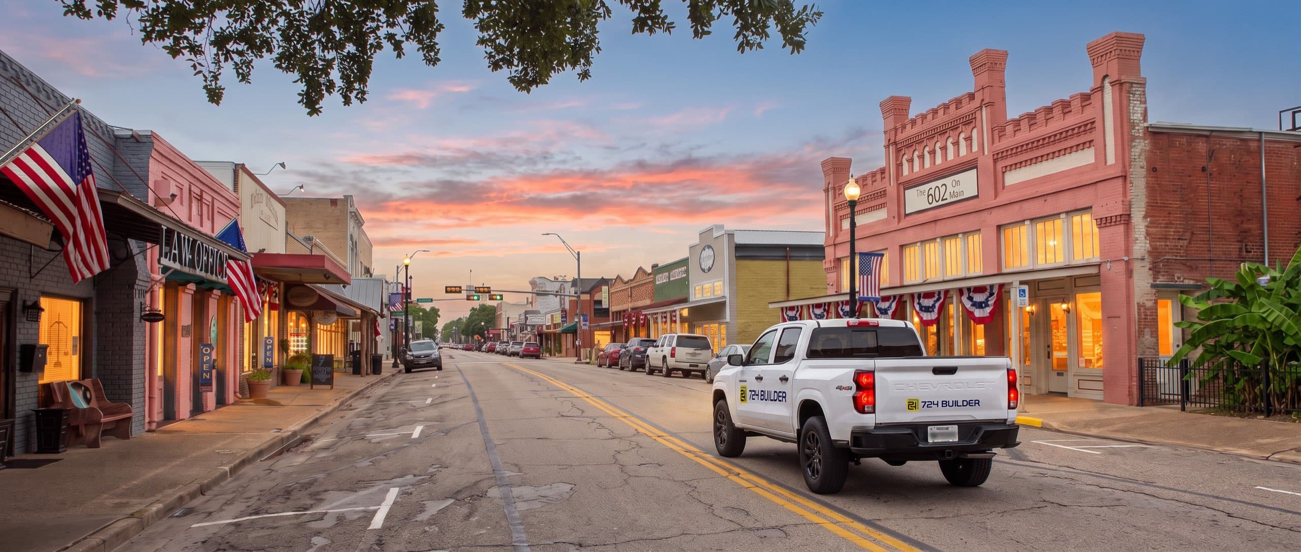 A white 724 Builder pickup truck drives away down the main street of downtown Bastrop at dusk, its tail lights lit. Historic brick and painted storefronts line both sides with their windows glowing warm, flags and bunting hanging from the canopies, and coral sunset cloud low over the rooflines.
