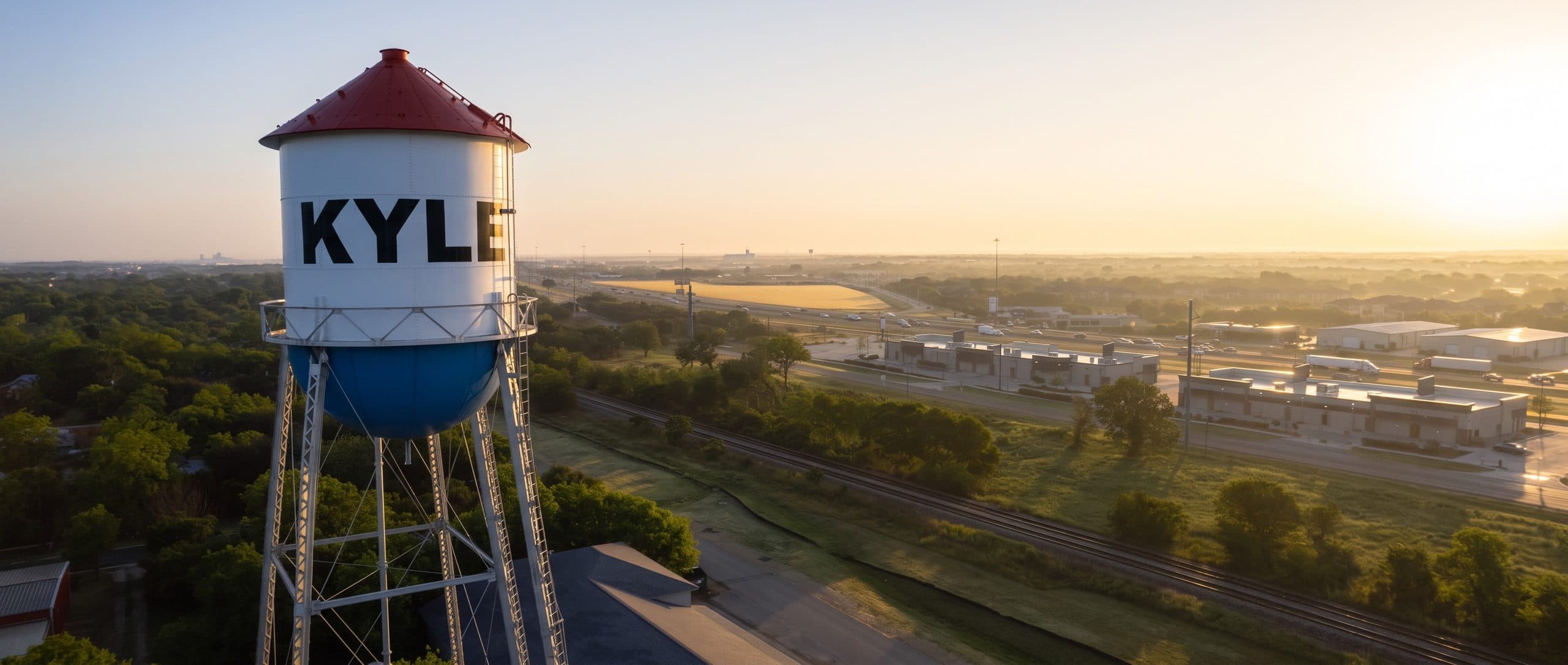 A steel water tower on tall lattice legs stands above Kyle, Texas at sunrise, its white tank painted with the town name in black block capitals under a deep red roof. Beyond it the land falls away to a rail line, the interstate and low commercial buildings, with morning haze on the horizon.