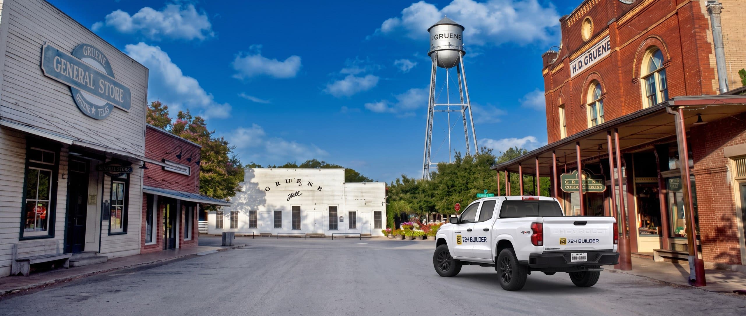 A white 724 Builder pickup truck drives away up the main street of the Gruene historic district in New Braunfels on a bright day. The Gruene water tower stands behind the rooflines with GRUENE lettered across its silver tank, the long white clapboard Gruene Hall sits at the end of the street, and the Gruene General Store's oval sign hangs from a timber porch on the left under a deep blue sky.
