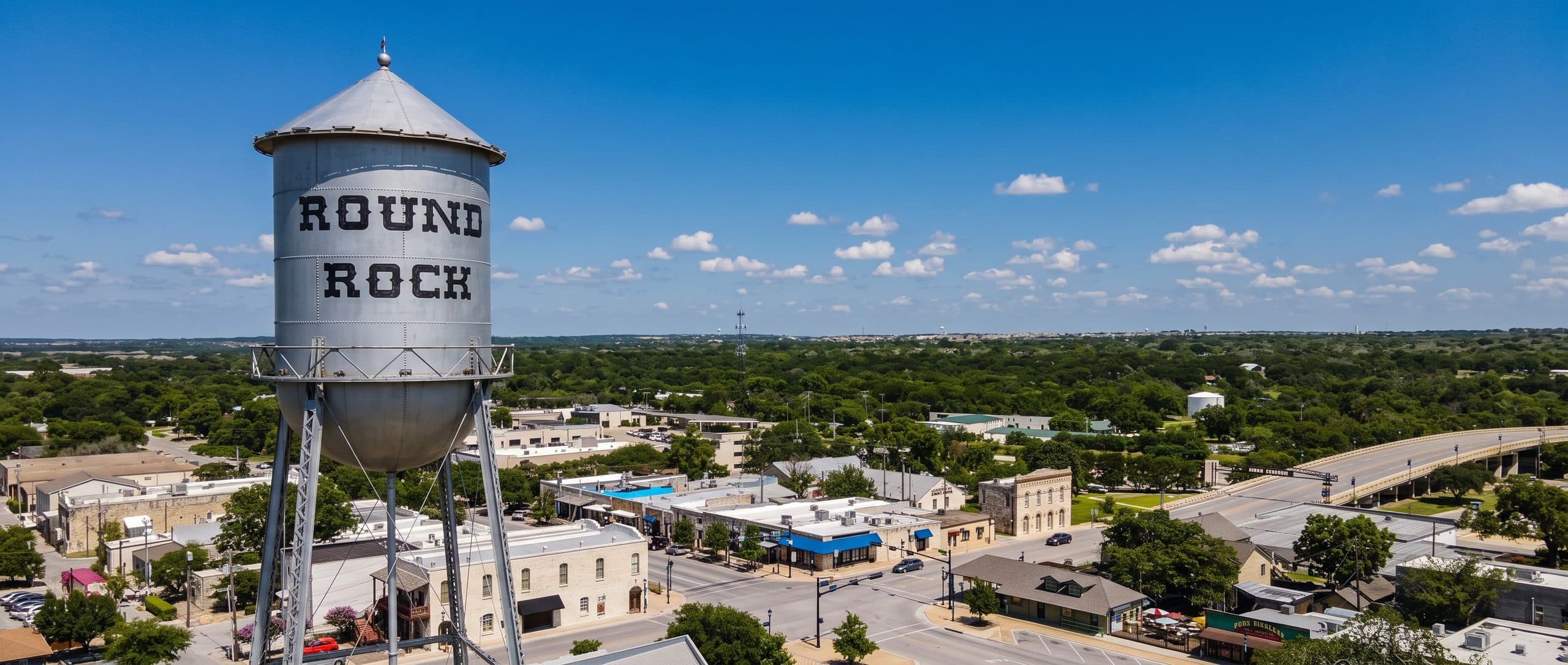 An aerial view of a tall galvanized steel water tower standing over downtown Round Rock, Texas, its tank painted with the town name in heavy black western lettering on two lines. Low brick and stone storefronts, a main street and mature shade trees spread out below under a deep blue sky with scattered cloud.
