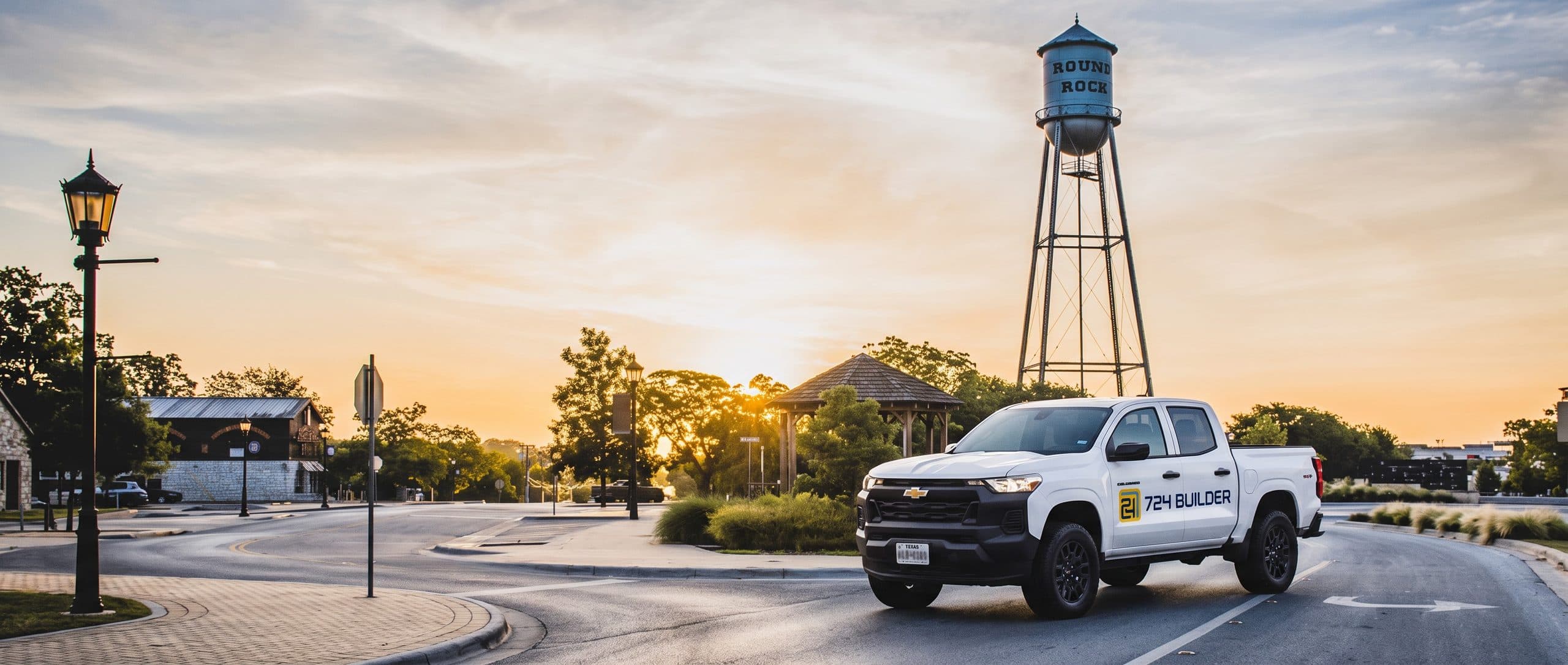 A white 724 Builder pickup truck turns onto a downtown street at sunrise, directly beneath a tall steel water tower whose blue-grey tank carries the words ROUND ROCK in weathered black lettering. A timber gazebo, low stone buildings and trees stand behind, with the sun low on the horizon.