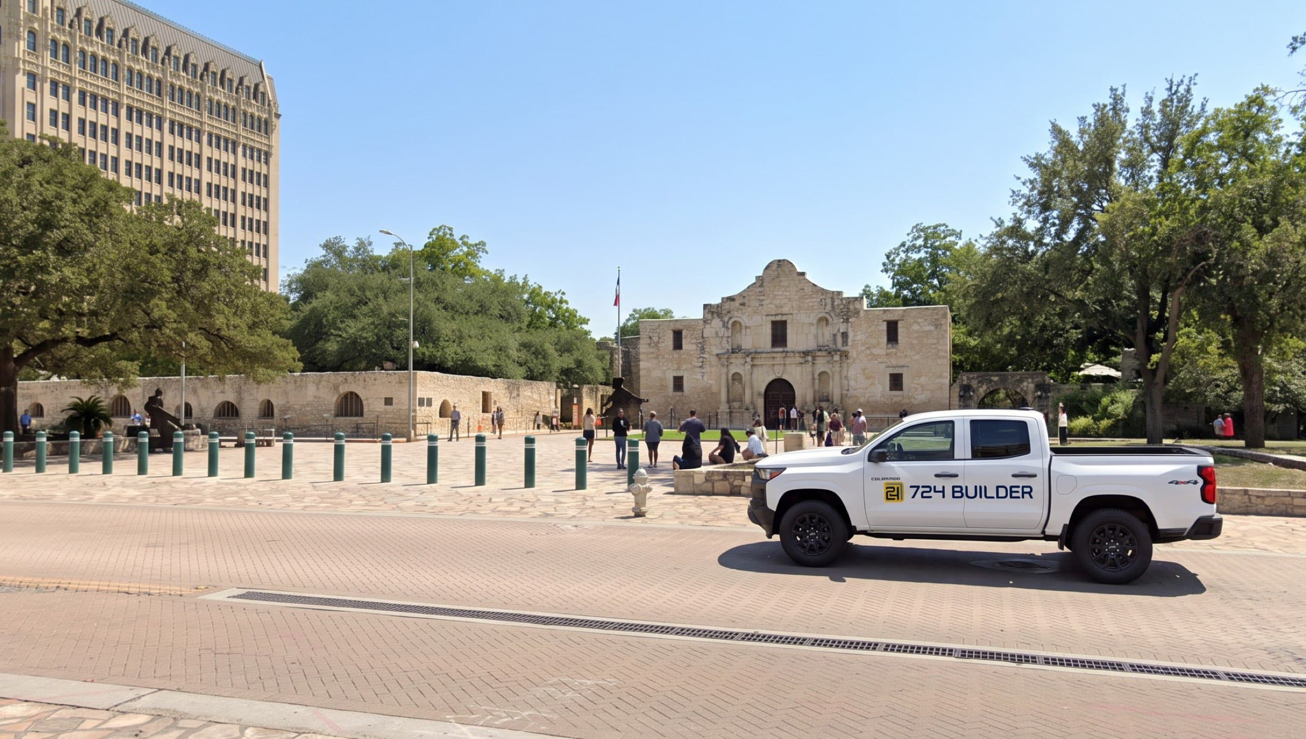 A white 724 Builder pickup truck parked on the street beside Alamo Plaza in San Antonio on a bright day. The limestone mission chapel with its curved gable stands across the plaza behind a low stone wall and a line of bollards, with live oaks around it and a tall historic tower building rising beyond the treeline.